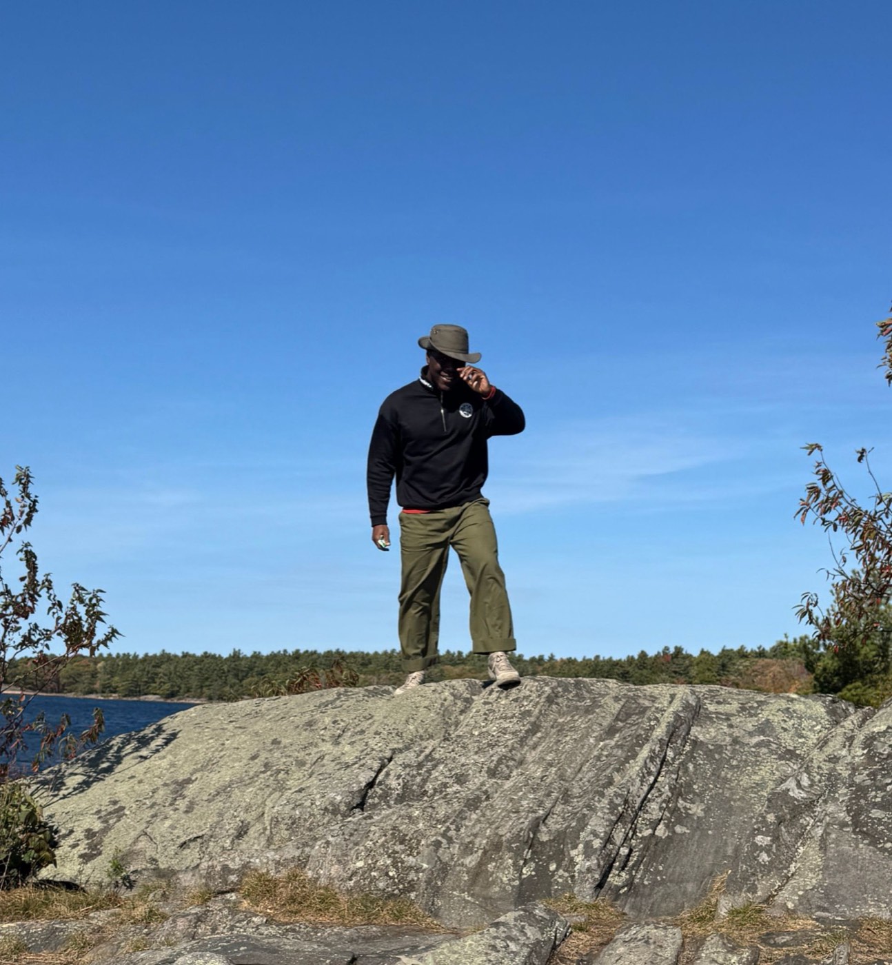 David on a granite outcrop by a lake, tipping his hat under a clear blue sky