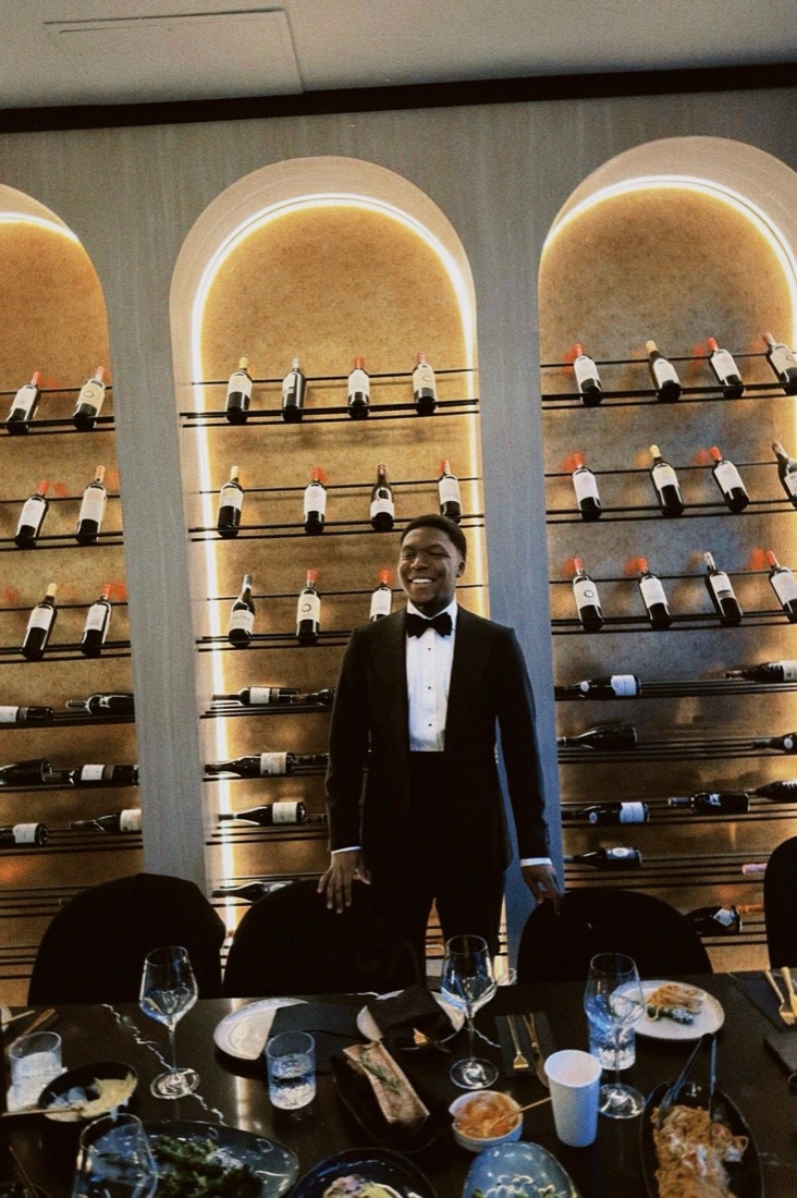David in black tie, smiling at a candlelit dinner before a wall of wine
